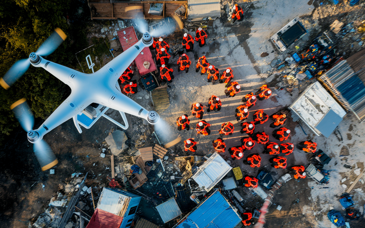 An aerial view of a drone flying over a disaster-stricken area, capturing real-time data for humanitarian efforts. Below, rescue teams are seen coordinating while assessing damaged structures and providing aid. The image highlights advanced technology's role in enhancing response efficiency, with vivid details of the terrain, logistics, and active teamwork below. Bright blue sky contrasting with the somber landscape emphasizes the urgency of the situation.