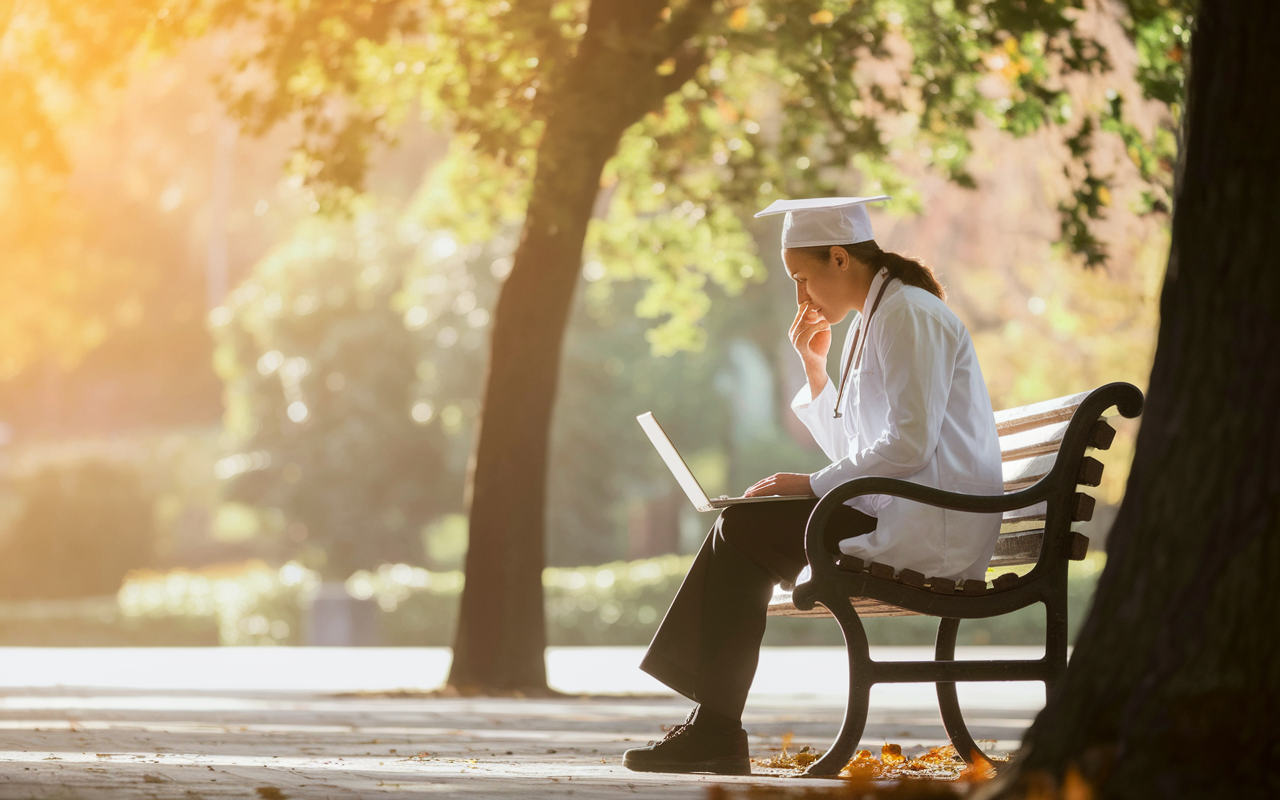 A pensive medical graduate alone in a serene park, sitting on a bench with their laptop open, holding a hand to their chin in contemplation, surrounded by trees and gentle autumn leaves. The golden hour sunlight casts a warm glow, suggesting introspection and the significance of their upcoming decision. The atmosphere feels tranquil yet charged with anticipation for the future.